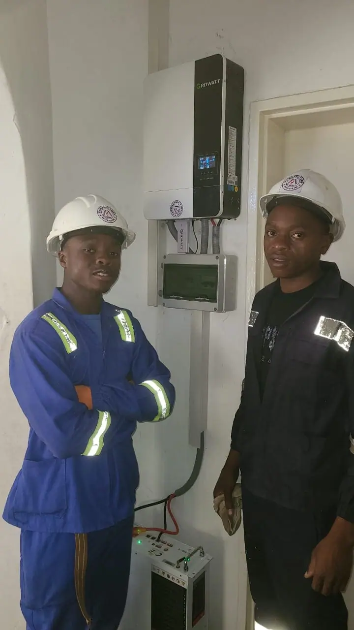 Two Pawafactory technicians in hard hats standing beside a wall-mounted Growatt inverter and battery